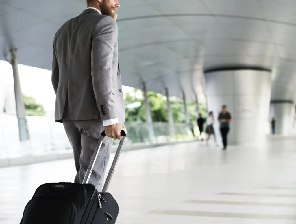 Nell'immagine si vede un uomo con abito elegante che trascina un trolley in un ambiente che ricorda quello di un aeroporto o una stazione. Sullo sfondo, si intravedono altre persone.