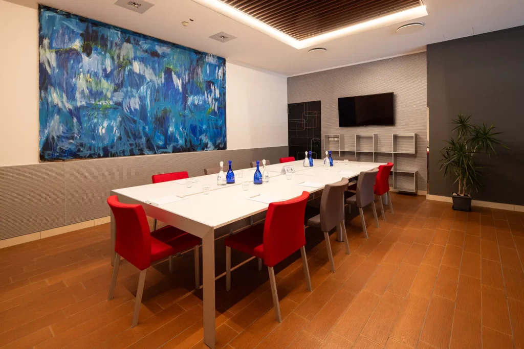 In the picture, the Callisto meeting room of the Hotel Galileo Padova: there is a table with chairs and, on the table, some notebooks with pens and some water bottles. Hanging on the wall in front of the table, there is a TV. On the other wall, a big abstract painting.