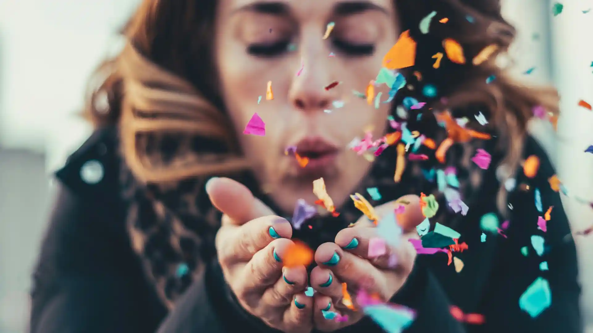 In the picture, a close-up of a woman blowing on some confetti she holds in her hands.